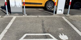 Ein Parkplatz für ein Elektroauto. Innerhalb der Parkplatz-Markierung ist ein Elektroauto abgebildet, was man an dem Stromkabel, das aus dem Fenster zeigt, sieht. Am Parkplatz befindet sich eine Ladesäule.