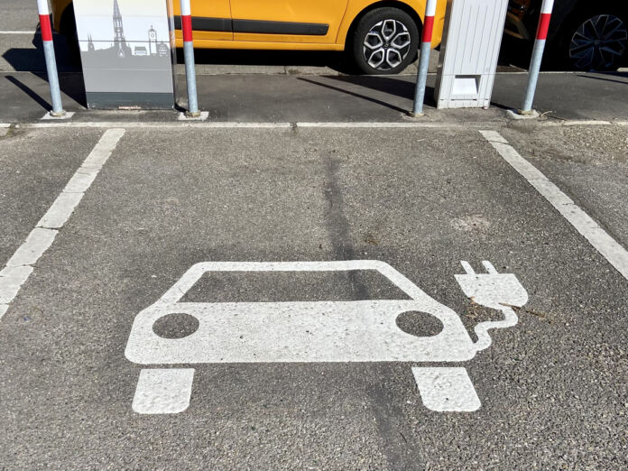 Ein Parkplatz für ein Elektroauto. Innerhalb der Parkplatz-Markierung ist ein Elektroauto abgebildet, was man an dem Stromkabel, das aus dem Fenster zeigt, sieht. Am Parkplatz befindet sich eine Ladesäule.