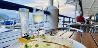 Auf einem Holztisch in einem Restaurant steht ein Teller mit Pommes Frites und Fisch. Dahinter steht ein kaltes Getränk im Glas und in einer Flasche. Das Restaurant befindet sich offensichtlich an Bord eines Schiffes, da man die Reling und das Wasser sieht.