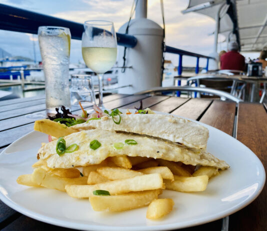 Auf einem Holztisch in einem Restaurant steht ein Teller mit Pommes Frites und Fisch. Dahinter steht ein kaltes Getränk im Glas und in einer Flasche. Das Restaurant befindet sich offensichtlich an Bord eines Schiffes, da man die Reling und das Wasser sieht.