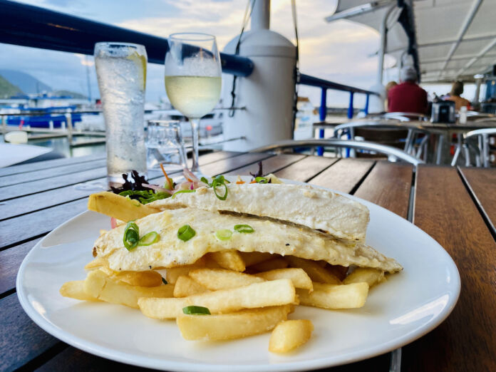 Auf einem Holztisch in einem Restaurant steht ein Teller mit Pommes Frites und Fisch. Dahinter steht ein kaltes Getränk im Glas und in einer Flasche. Das Restaurant befindet sich offensichtlich an Bord eines Schiffes, da man die Reling und das Wasser sieht.