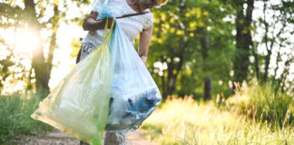 Eine ältere Frau in weißem Shirt und Sandalen trägt Plastiktüten in der Hand. Sie läuft auf einem Sandweg im Wald und hebt Plastik-Müll auf. Sie geht gegen Umweltverschmutzung vor.