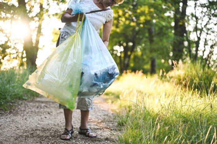 Eine ältere Frau in weißem Shirt und Sandalen trägt Plastiktüten in der Hand. Sie läuft auf einem Sandweg im Wald und hebt Plastik-Müll auf. Sie geht gegen Umweltverschmutzung vor.