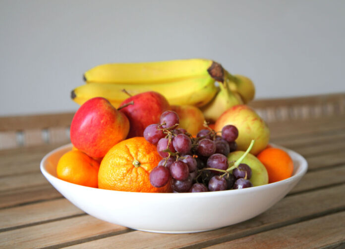 Eine Schale mit frischem Obst und Früchten steht auf einem Tisch. Darunter befinden sich Bananen, Äpfel, Orangen und Weintrauben.