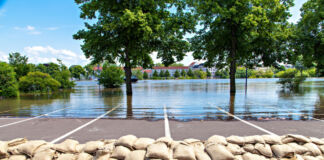 Nach langen Regenfällen gibt es Hochwasser auf einem Parkplatz. Im Hintergrund stehen die Bäume und Häuser unter Wasser. Deshalb haben einige Menschen und Bürger Sandsäcke aufeinander gestapelt.