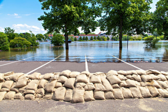 Nach langen Regenfällen gibt es Hochwasser auf einem Parkplatz. Im Hintergrund stehen die Bäume und Häuser unter Wasser. Deshalb haben einige Menschen und Bürger Sandsäcke aufeinander gestapelt.