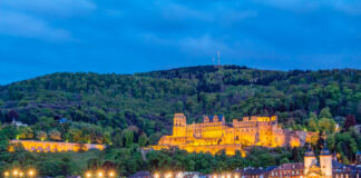 Ein Panoramafoto, auf dem man eine beleuchtete Brücke sieht. Die Steinbrücke steht vor einem bewaldeten Hügel und einem wunderschönen Schloss. Die Schlossruine befindet sich über der Altstadt. Ein beliebter Ort bei Touristen.