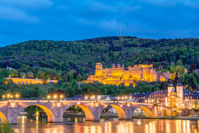 Ein Panoramafoto, auf dem man eine beleuchtete Brücke sieht. Die Steinbrücke steht vor einem bewaldeten Hügel und einem wunderschönen Schloss. Die Schlossruine befindet sich über der Altstadt. Ein beliebter Ort bei Touristen.