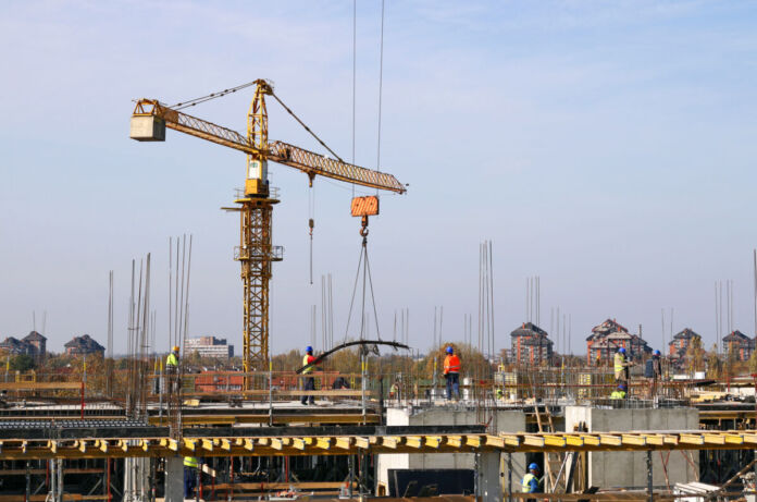 Eine große Baustelle, auf der ein Kran im Einsatz ist. Unten arbeiten die Bauarbeiter und stellen die Gerüste auf. Ein neues großes Gebäude oder ein Hochhaus aus Betonelementen entsteht.