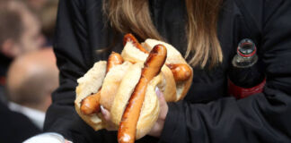 Eine Frau trägt eine Cola unter dem Arm. In der anderen Hand hält sie ein Bier und drei Brötchen mit einer Bratwurst. Die Frau befindet sich offensichtlich in einem Fußballstadion.