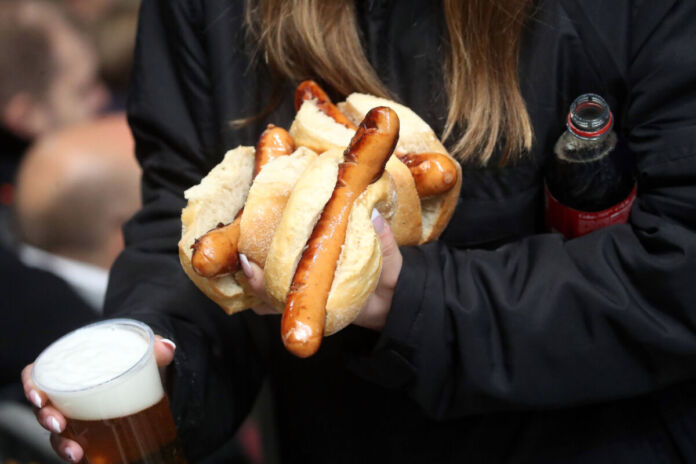 Eine Frau trägt eine Cola unter dem Arm. In der anderen Hand hält sie ein Bier und drei Brötchen mit einer Bratwurst. Die Frau befindet sich offensichtlich in einem Fußballstadion.