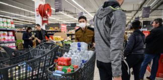 In einem Discounter oder Supermarkt haben einige Kunden ihre Einkaufswagen bis oben hin mit Getränken vollgepackt. Im Hintergrund schauen sich Kunden einige Artikel an.