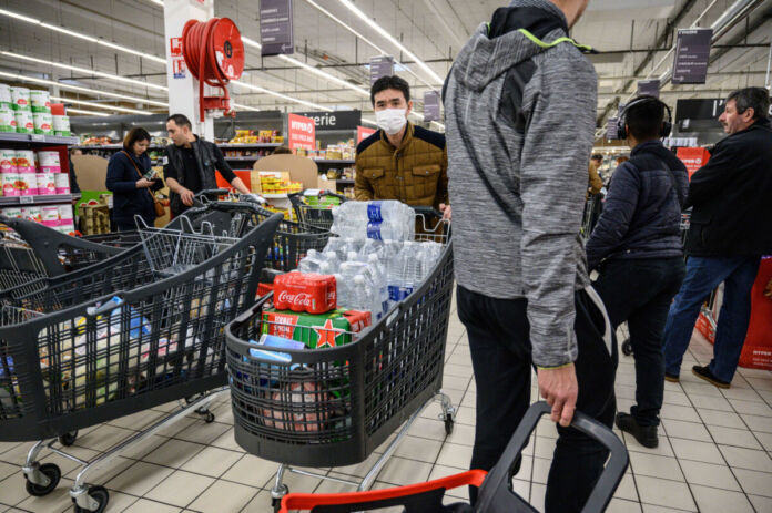 In einem Discounter oder Supermarkt haben einige Kunden ihre Einkaufswagen bis oben hin mit Getränken vollgepackt. Im Hintergrund schauen sich Kunden einige Artikel an.