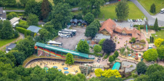 Aus der Luftperspektive sieht man einen Freizeitpark mit Besuchern und seinen vielen Attraktionen. Busse parken auf dem Parkplatz neben dem Park. Rutschen, Karussells und ein großer Spielplatz sind zu sehen.