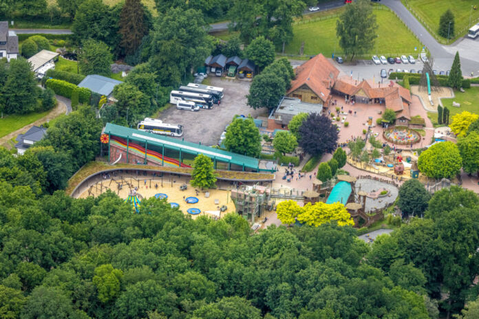 Aus der Luftperspektive sieht man einen Freizeitpark mit Besuchern und seinen vielen Attraktionen. Busse parken auf dem Parkplatz neben dem Park. Rutschen, Karussells und ein großer Spielplatz sind zu sehen.