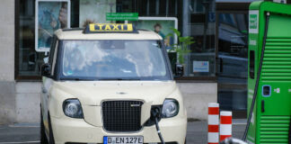 Ein Elektro-Taxi ist an einer Ladesäule angeschlossen, um seine Batterie aufzuladen. Der Stromer hat die klassische hell-elfenbeinfarbenen Farbe und ist Teil einer E-Taxi-Flotte. Die Schnellladestation ist grün.