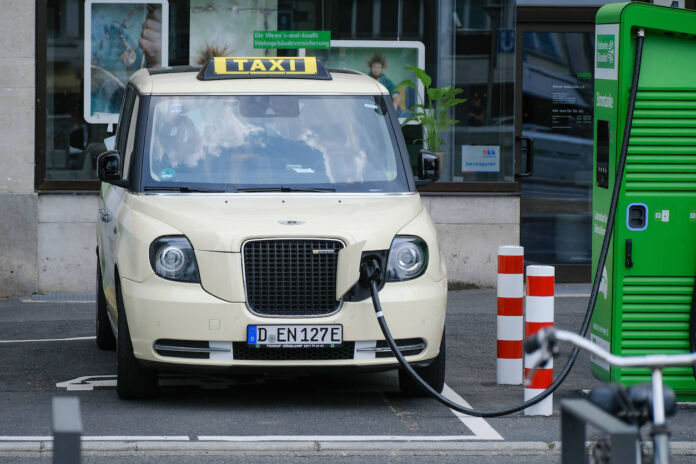 Ein Elektro-Taxi ist an einer Ladesäule angeschlossen, um seine Batterie aufzuladen. Der Stromer hat die klassische hell-elfenbeinfarbenen Farbe und ist Teil einer E-Taxi-Flotte. Die Schnellladestation ist grün.