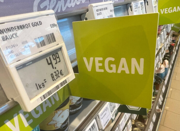 Im Regal eines Supermarktes oder eines Discounters gibt es eine große Auswahl an verschiedenen veganen Produkten. Ein grünes großes Schild mit der Aufschrift "Vegan" weist darauf hin. Man sieht ein Preisschild mit dem Hinweis, dass es sich hier um veganes Brot handelt.