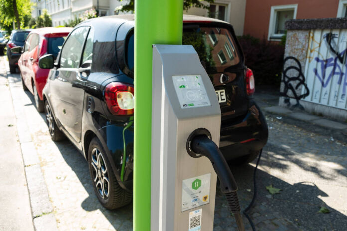 Ein kleines schwarzes E-Auto lädt an einem grünen Laternenmast seine Batterie per Ladekabel auf. Der Smart steht in einer Reihe mit anderen Autos am Straßenrand geparkt.