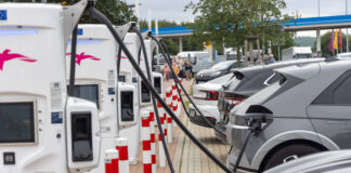 Elektroautos stauen sich an einer Autobahnraststätte, um mit Strom geladen zu werden. Die E-Autos stehen in einer Reihe und sind an die weißen Ladesäulen angeschlossen. Im Hintergrund dieser Stromtankstelle steht eine normale Tankstelle.