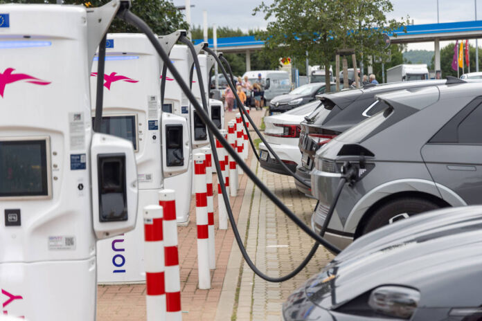 Elektroautos stauen sich an einer Autobahnraststätte, um mit Strom geladen zu werden. Die E-Autos stehen in einer Reihe und sind an die weißen Ladesäulen angeschlossen. Im Hintergrund dieser Stromtankstelle steht eine normale Tankstelle.