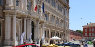 Drei sehr luxuriöse Autos stehen bei gutem Wetter aufgereiht auf einem Marktplatz. Es handelt sich um Sportwagen – ein roter, goldener und silberner. Sie stehen auf kleinen Bühnen wie für eine Ausstellung oder Parade.