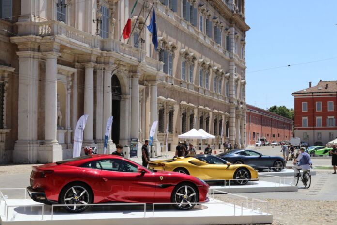 Drei sehr luxuriöse Autos stehen bei gutem Wetter aufgereiht auf einem Marktplatz. Es handelt sich um Sportwagen – ein roter, goldener und silberner. Sie stehen auf kleinen Bühnen wie für eine Ausstellung oder Parade.