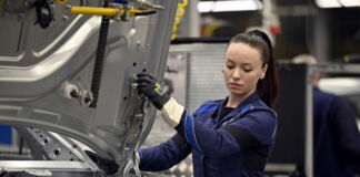 Eine Mitarbeiterin in der Halle eines Autobauers hält in ihren Händen gerade die Motorhaube und führt sie in die Vorrichtung zur Befestigung ein. Man sieht den Rohbau des Fahrzeugs im Vordergrund. Im Hintergrund sitzt ein älterer Mitarbeiter vor einem Bildschirm.