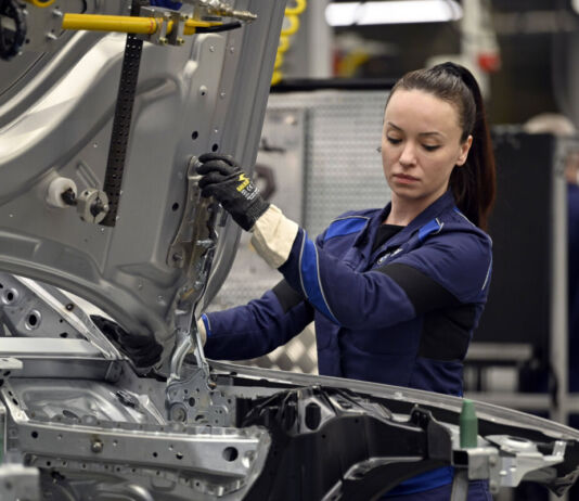 Eine Mitarbeiterin in der Halle eines Autobauers hält in ihren Händen gerade die Motorhaube und führt sie in die Vorrichtung zur Befestigung ein. Man sieht den Rohbau des Fahrzeugs im Vordergrund. Im Hintergrund sitzt ein älterer Mitarbeiter vor einem Bildschirm.