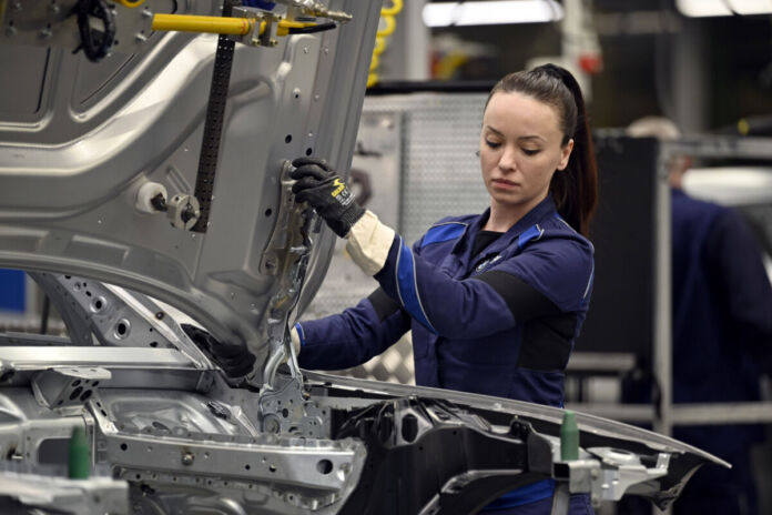 Eine Mitarbeiterin in der Halle eines Autobauers hält in ihren Händen gerade die Motorhaube und führt sie in die Vorrichtung zur Befestigung ein. Man sieht den Rohbau des Fahrzeugs im Vordergrund. Im Hintergrund sitzt ein älterer Mitarbeiter vor einem Bildschirm.