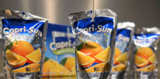 Mehrere Capri-Sun-Fruchtsaftgetränke der Sorte Orange in Kunststoffbeuteln auf einem Tisch. Im Hintergrund ist neben zwei der Getränke eine Kartonverpackung für die einzelnen Säfte zu sehen.