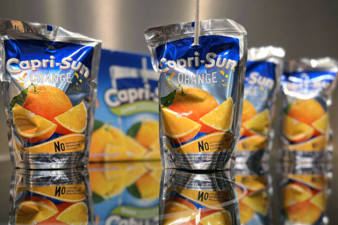 Mehrere Capri-Sun-Fruchtsaftgetränke der Sorte Orange in Kunststoffbeuteln auf einem Tisch. Im Hintergrund ist neben zwei der Getränke eine Kartonverpackung für die einzelnen Säfte zu sehen.