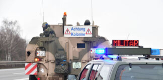 Ein Militärfahrzeug mit der dem Warnschild "Achtung Kolonne", in dem sich Soldaten befinden, fährt gerade auf die Autobahn. Hinter ihm fährt ein weiteres Fahrzeug mit Blaulicht und der Warnung Militär.