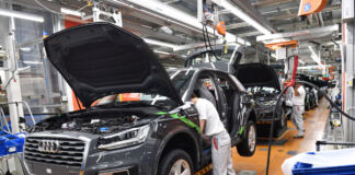 Auf einer Fertigungsstraße in einer Fertigungshalle in einem AUDI-Werk stehen viele schwarze Fahrzeuge in einer Reihe. Mehrere Arbeiter montieren etwas am Fahrzeug.