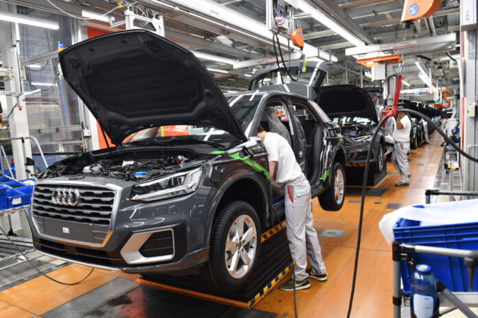 Auf einer Fertigungsstraße in einer Fertigungshalle in einem AUDI-Werk stehen viele schwarze Fahrzeuge in einer Reihe. Mehrere Arbeiter montieren etwas am Fahrzeug.