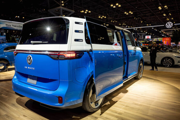 Ein blauer Elektrobus von VW wird auf einer Autoshow präsentiert. Er steht auf einem beleuchteten Podium, umgeben von Interessierten sowie anderen Autos und wird von hellen Lichtern angestrahlt.