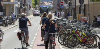 Mehrere Fahrradfahrer fahren auf der Straße und auf einer Fahrradstraße. Neben der Straße stehen viele Fahrräder nebeneinander. Es handelt sich offensichtlich um eine Fahrradstadt.