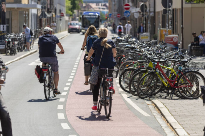 Mehrere Fahrradfahrer fahren auf der Straße und auf einer Fahrradstraße. Neben der Straße stehen viele Fahrräder nebeneinander. Es handelt sich offensichtlich um eine Fahrradstadt.