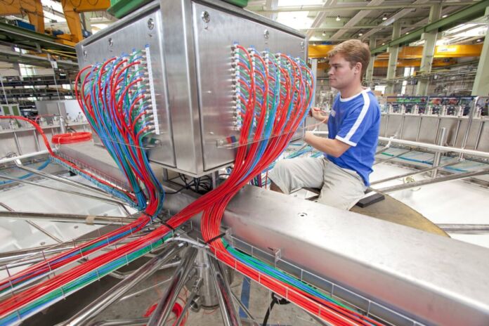 Inmitten eines Werkes steht ein Behälter aus Edelstahl oder Aluminium, in welchem blaue und rote Kabel stecken. Die farbigen Stromkabel laufen aus vielen Richtungen zentral zusammen. Neben der Anlage sitzt ein junger blonder Mann mit blauem Pulli und ist mit der Elektromontage beschäftigt.