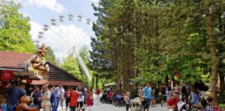 Männer, Frauen und Familien laufen entspannt bei gutem Wetter durch einen Freizeitpark. Im Park gibt es ein Riesenrad, Achterbahnen und viele weitere Attraktionen.