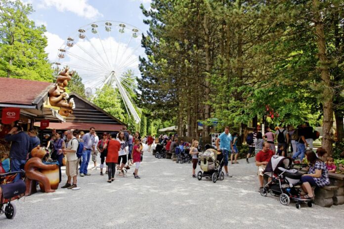 Männer, Frauen und Familien laufen entspannt bei gutem Wetter durch einen Freizeitpark. Im Park gibt es ein Riesenrad, Achterbahnen und viele weitere Attraktionen.