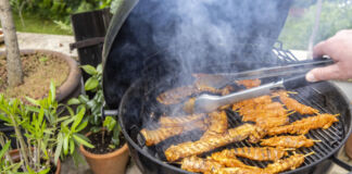 Eine Person hält eine Grillzange in der Hand und wendet das Grillgut auf dem Grill. Es handelt sich um Fleisch, vegetarische und vegane Speisen, die über dem Feuer gegrillt werden.