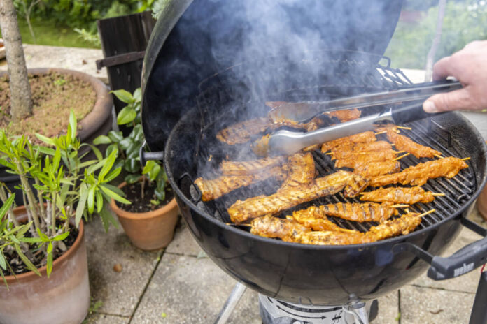 Eine Person hält eine Grillzange in der Hand und wendet das Grillgut auf dem Grill. Es handelt sich um Fleisch, vegetarische und vegane Speisen, die über dem Feuer gegrillt werden.