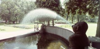 In einem Park steht ein Brunnen, bei dem es sich um eine Statue handelt, die Wasser speit. Die Parkanlage ist grün und ein wunderschönes Ausflugsziel für Familien. Im Park stehen zahlreiche Bäume.