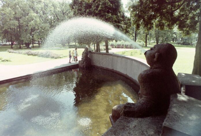 In einem Park steht ein Brunnen, bei dem es sich um eine Statue handelt, die Wasser speit. Die Parkanlage ist grün und ein wunderschönes Ausflugsziel für Familien. Im Park stehen zahlreiche Bäume.