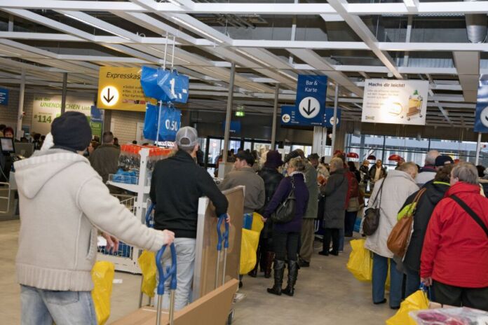 In einem IKEA-Markt stehen viele Menschen an verschiedenen Kassen in einer langen Schlange und warten ungeduldig darauf, dass ihre ausgesuchten Waren abkassiert werden.