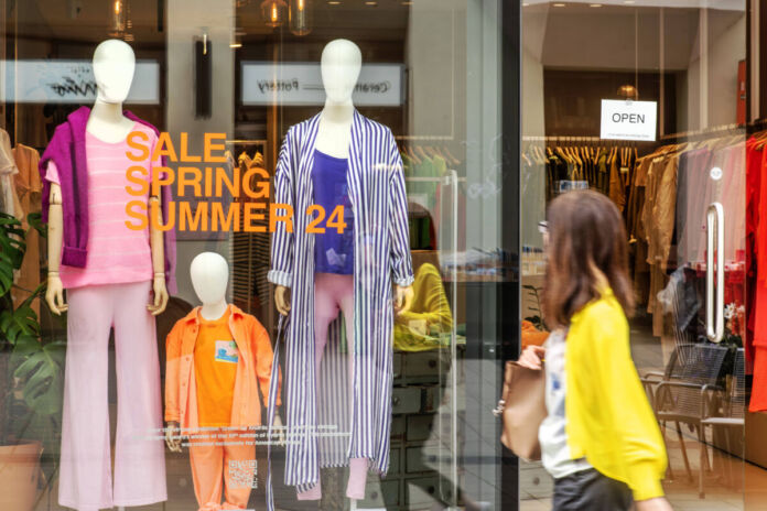 Im Schaufenster der Filiale einer großen Mode-kette stehen Figuren mit Anziehsachen, welche stark reduziert sind. Eine Frau läuft am Schaufenster des Modehauses vorbei.