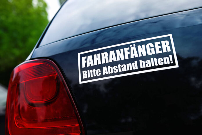 Eine Nahaufnahme von einem Heck eines schwarzen Pkw. Es ist ein weißer Aufkleber angebracht, auf dem steht "Fahranfänger – Bitte Abstand halten!"