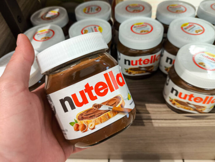 Eine Hand hält ein Glas Nutella. Dahinter stehen viele Gläser der Nuss-Nougat-Creme aufgereiht, wahrscheinlich in einem Regal. Es handelt sich um das klassische Design der Schokocreme.