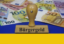 Ein Stempel mit dem Aufdruck "Bürgergeld" liegt auf einer ukrainischen Flagge. Hier liegt auch eine Menge Bargeld, das den staatlichen Zuschuss für Flüchtlinge symbolisiert. Es sind 5-Euro, 10-Euro, 20-Euro, 50-Euro-, 100-Euro, 200-Euro und ein 500-Euro-Schein.